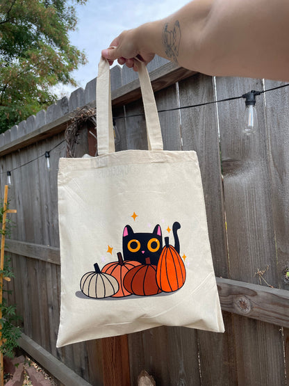 Tote bag with a black cat and pumpkins design held by a person outdoors.